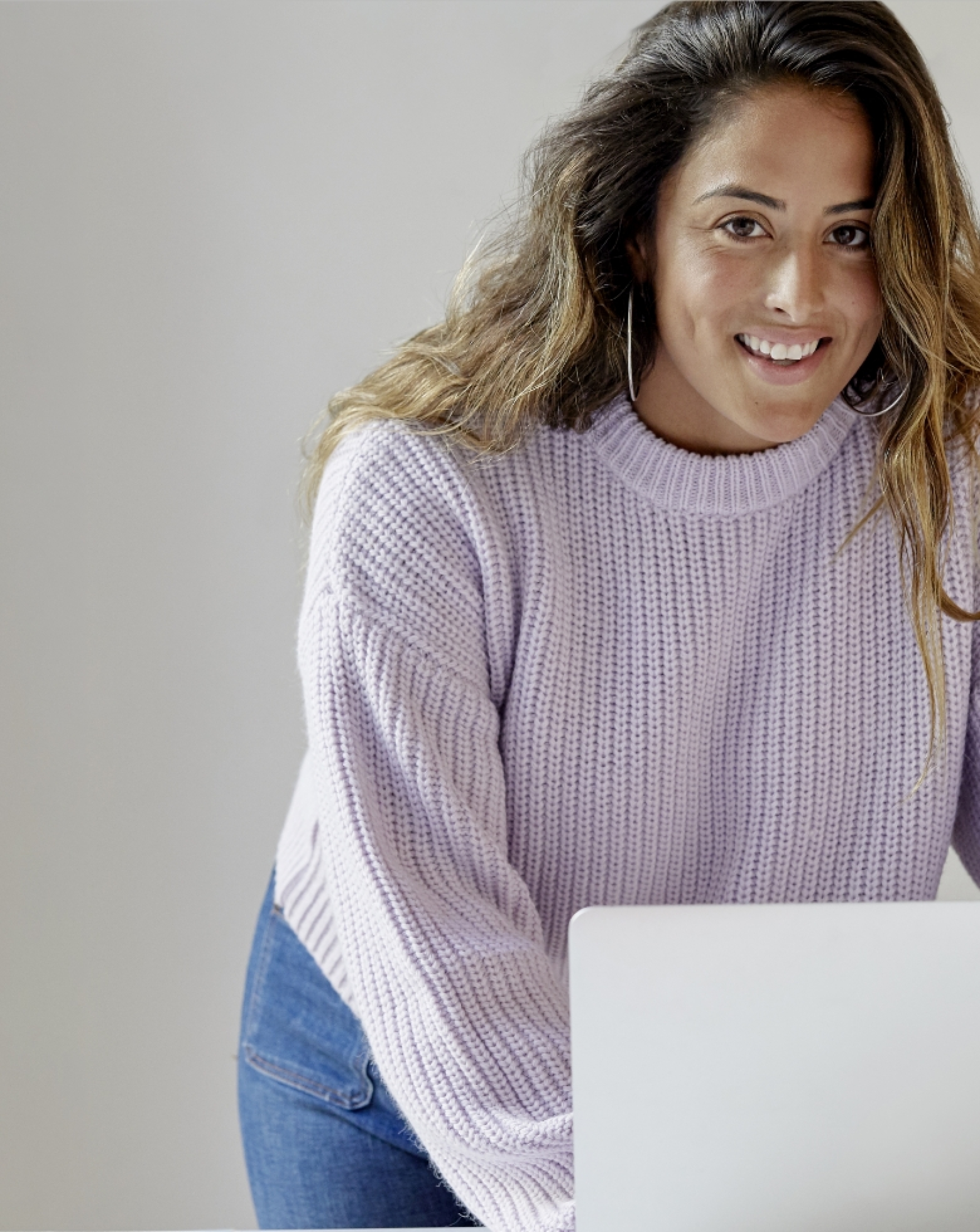 Young friendly looking woman in lilac jumper in working environment smiling at the camera with a laptop in front of her.