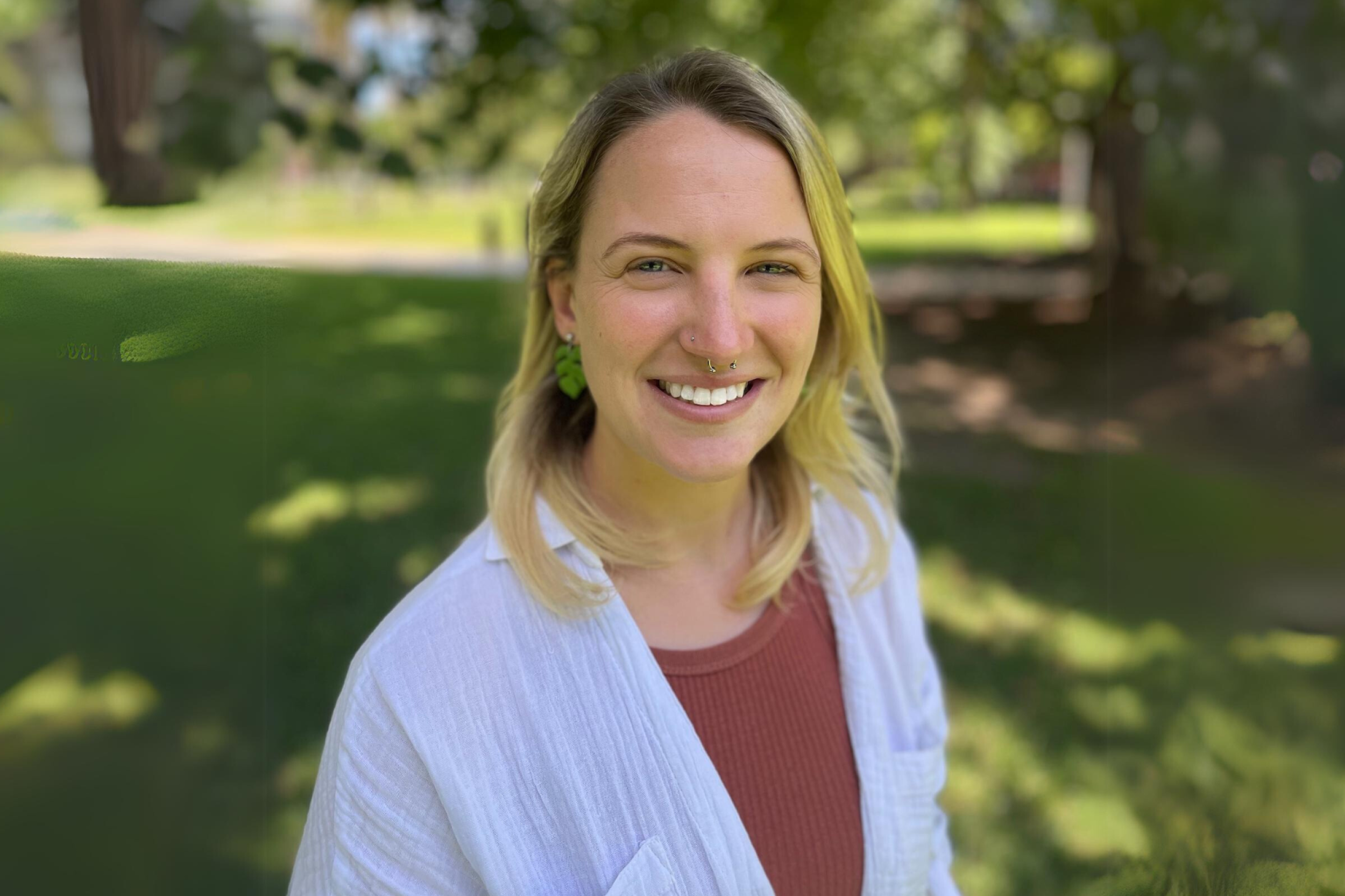 A blonde woman in a park smiles at the camera.