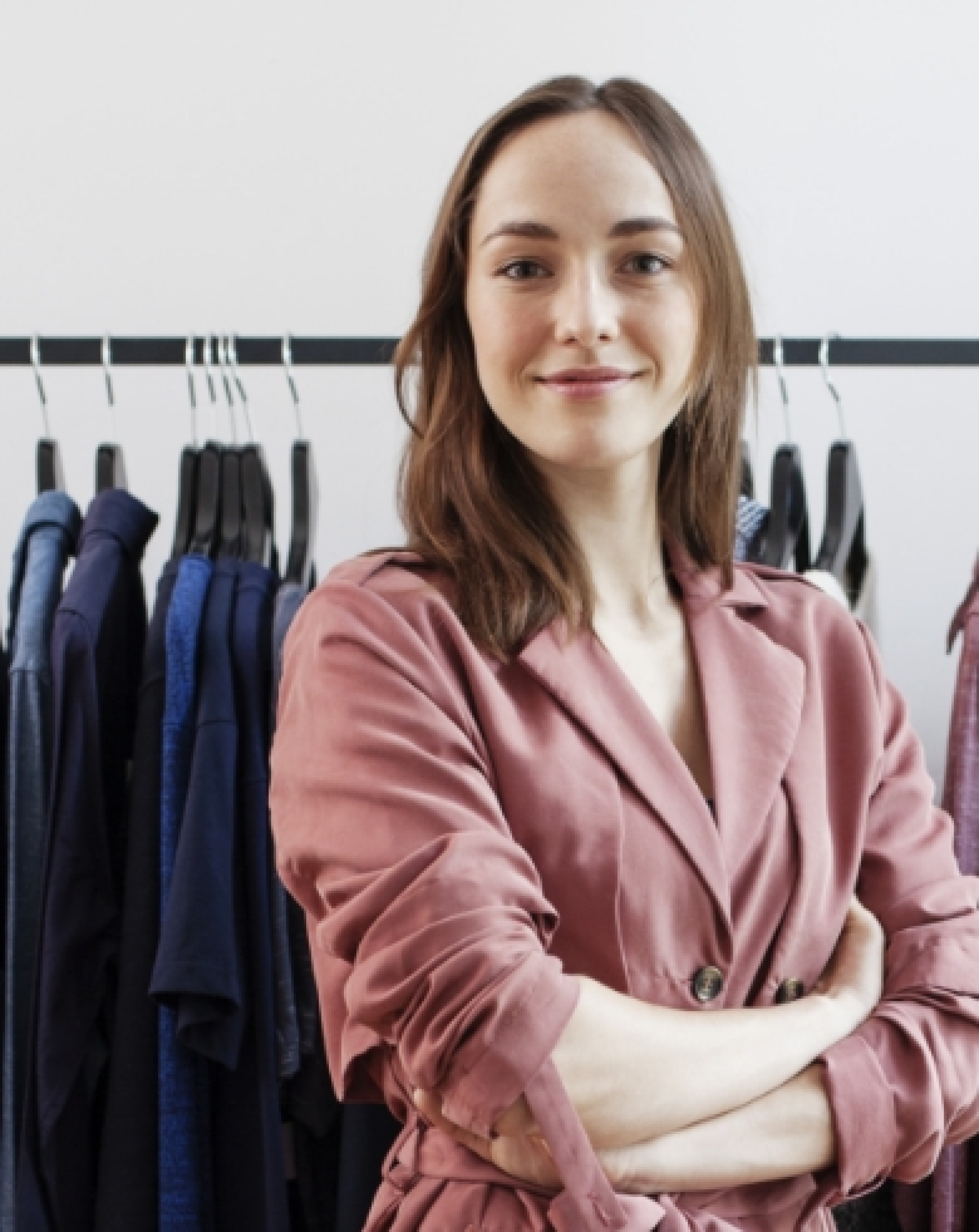 Young business woman looking proud and smiling at the camera while standing in front of a rack of clothing in her own store.