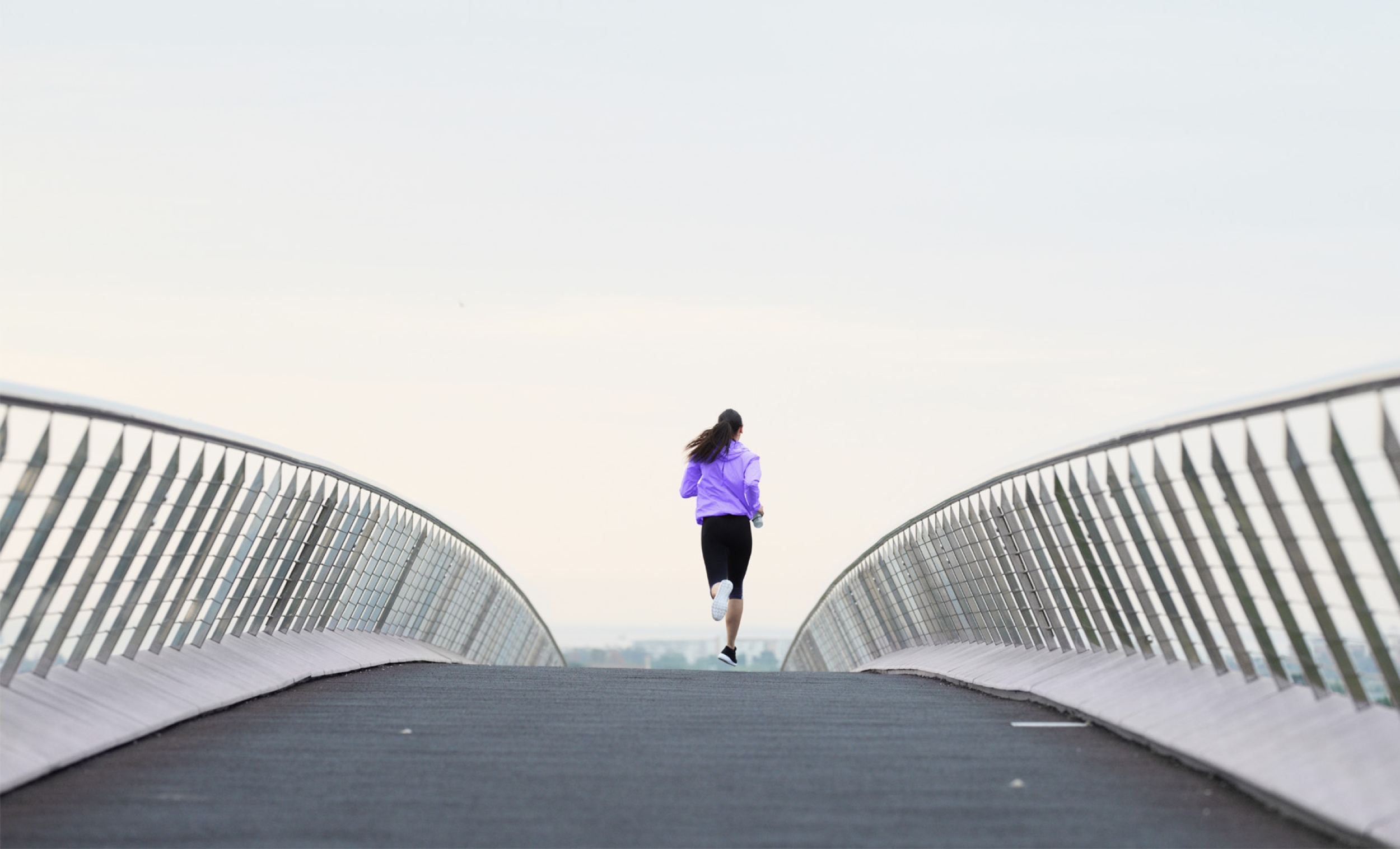 Young athletic woman seen from the back; she is wearing a purple jacket and is running from the viewer over a bridge.