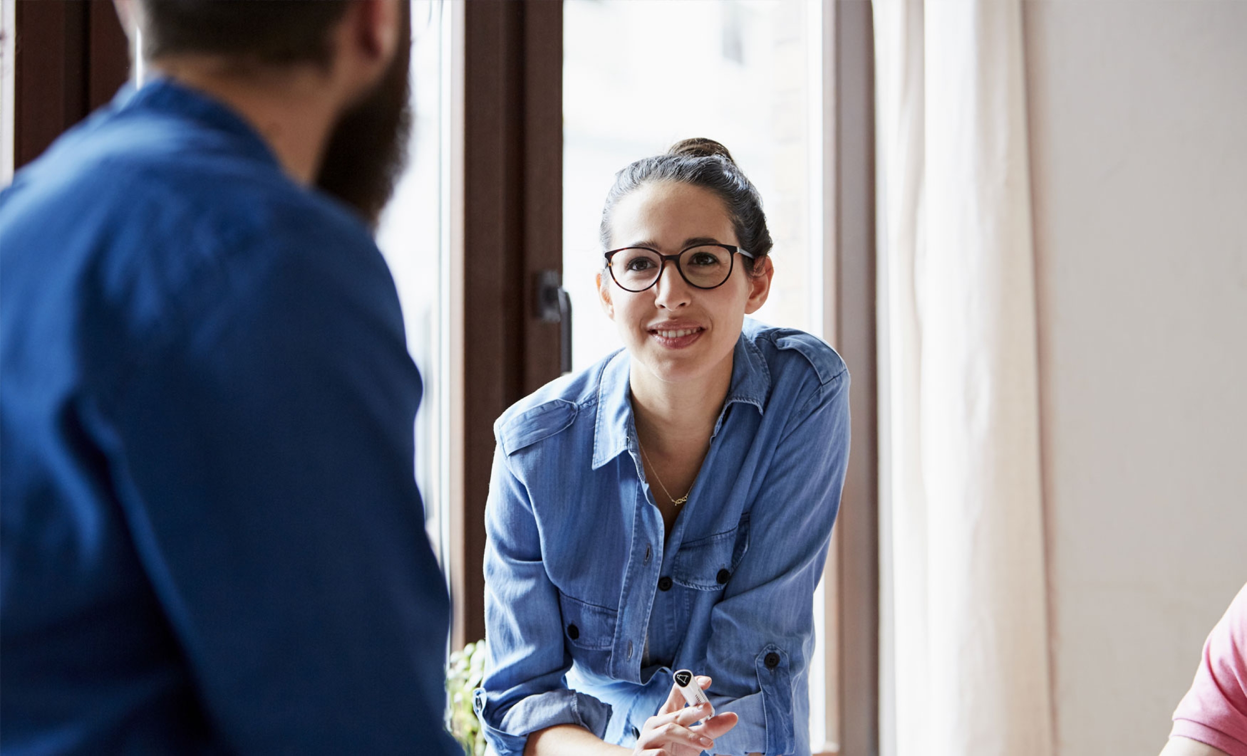 Office environment showing colleagues at work – a young woman dressed in a denim shirt and glasses is holding a pen and smiling at her colleague.