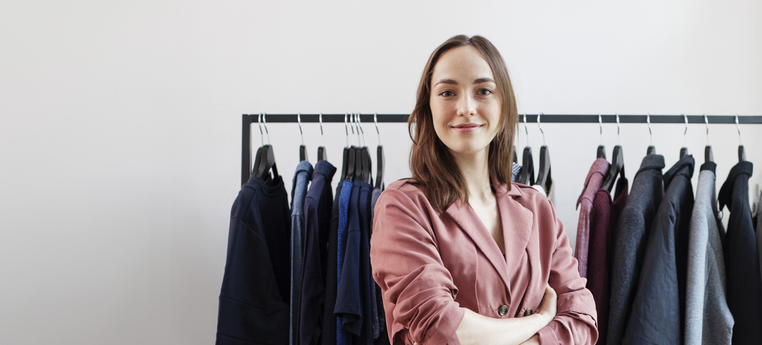 Young business woman looking proud and smiling at the camera while standing in front of a rack of clothing in her own store.