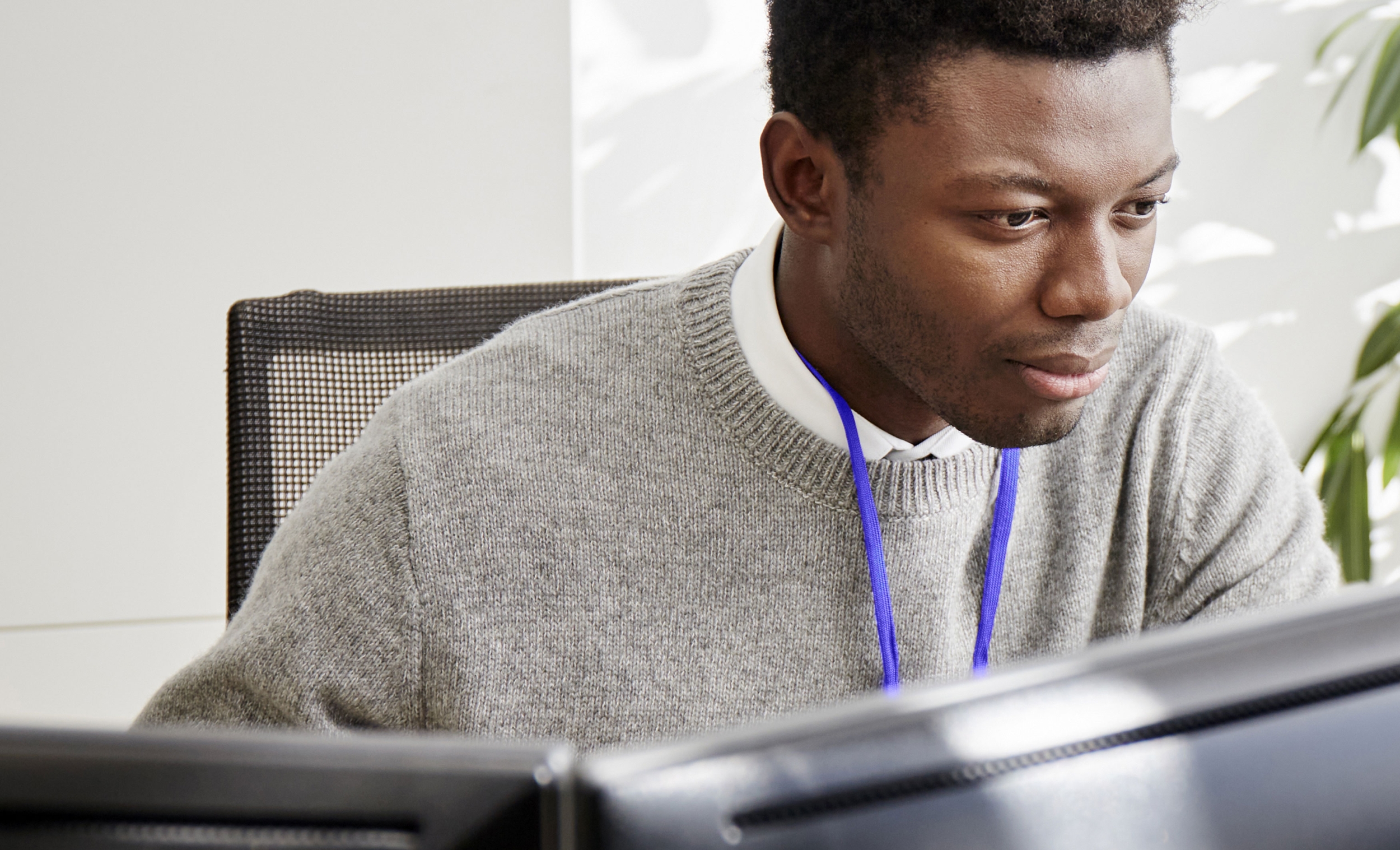 Young black business man wearing a grey jumper and a purple keycord. He is sitting in an office environment and is focused at a double monitor screen in front of him.