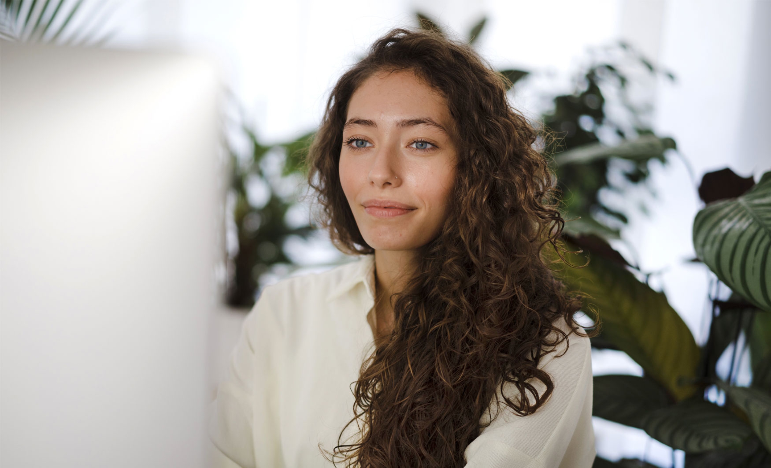 Young woman with brown hair in an office environment smiling and looking at a monitor while at work.