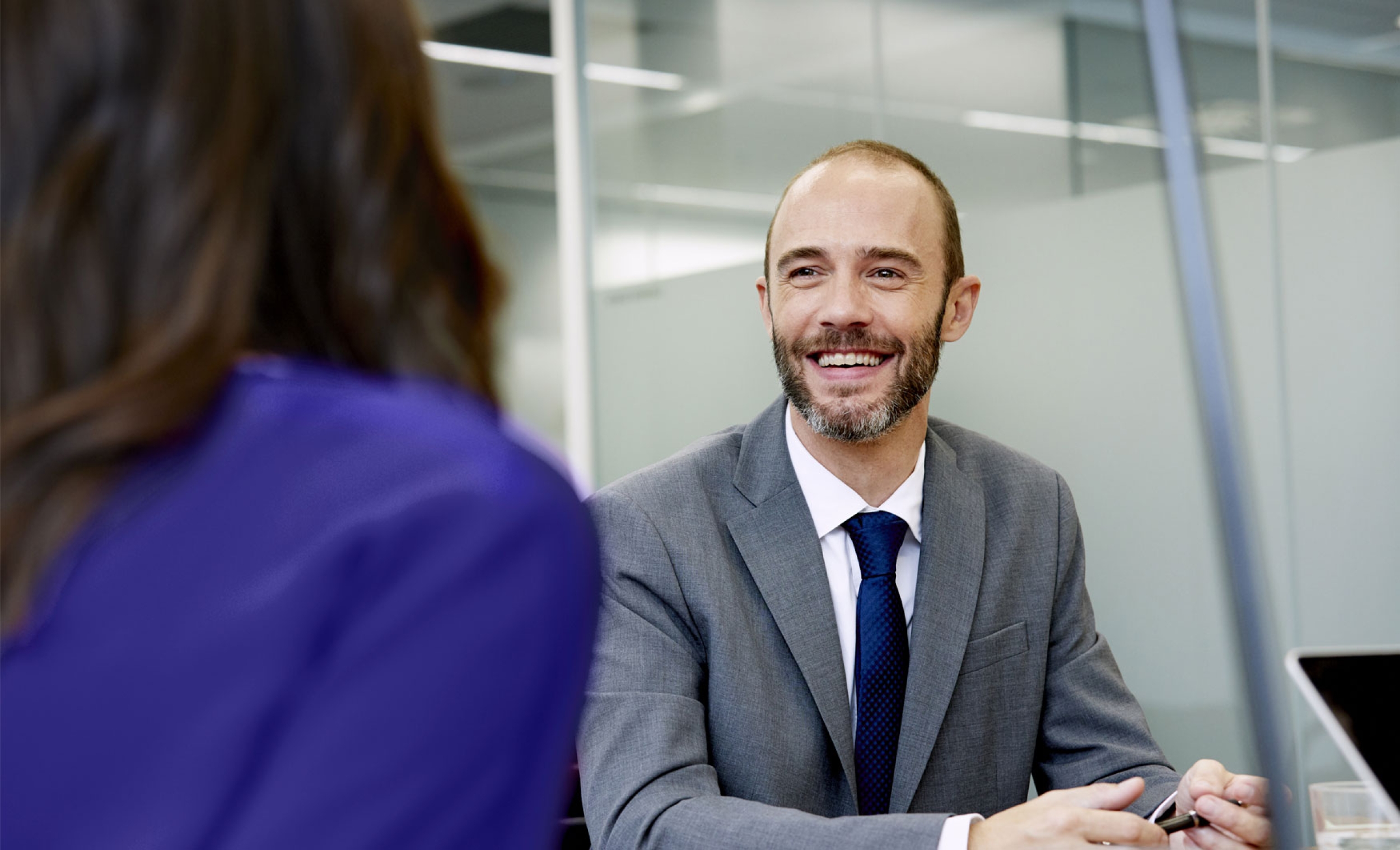 A middle-aged business professional is wearing a grey suit and blue tie while smiling at his colleague in an office environment.