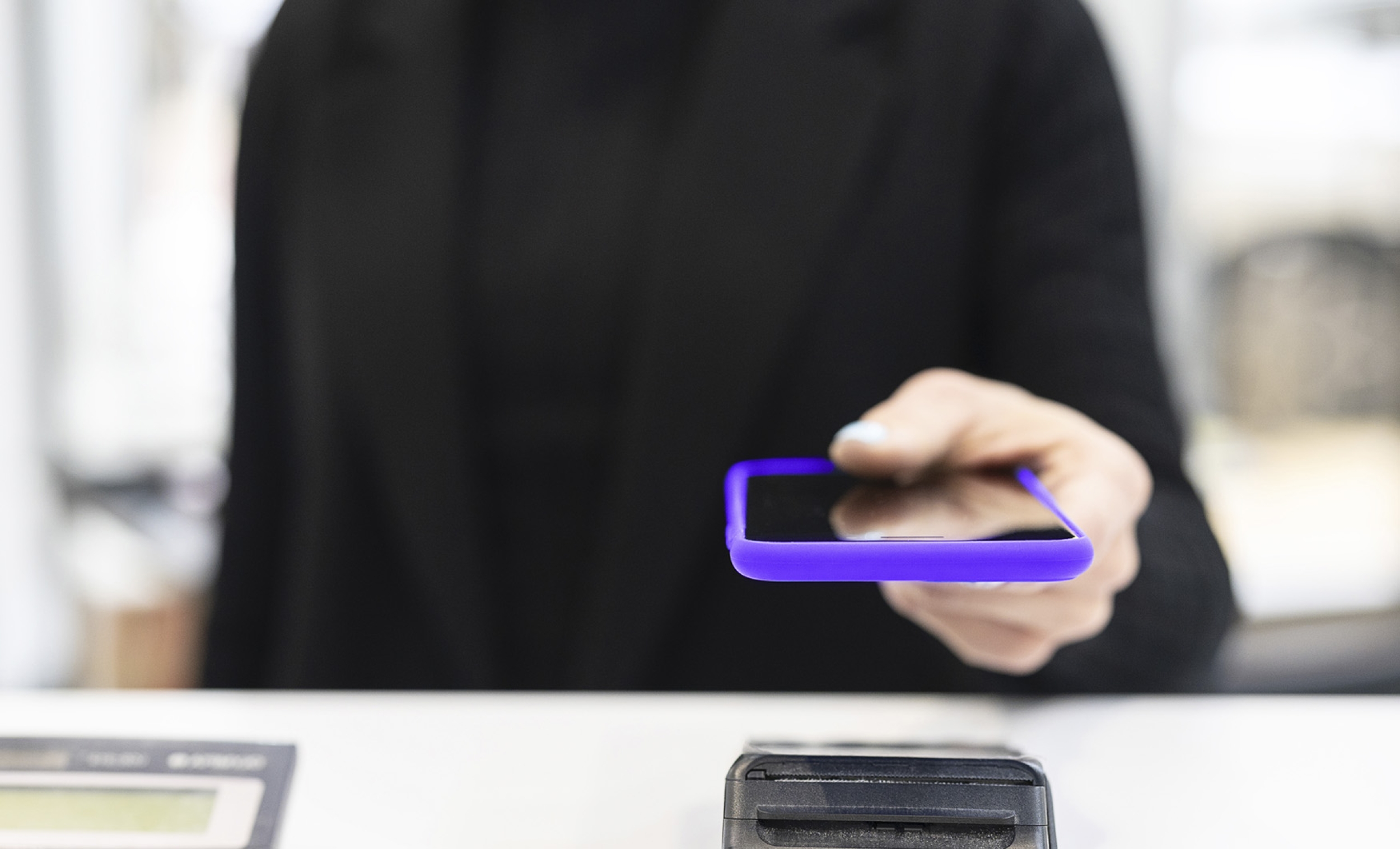Close-up shot of a woman holding a phone with purple cover to make a payment on a payments device in a store.