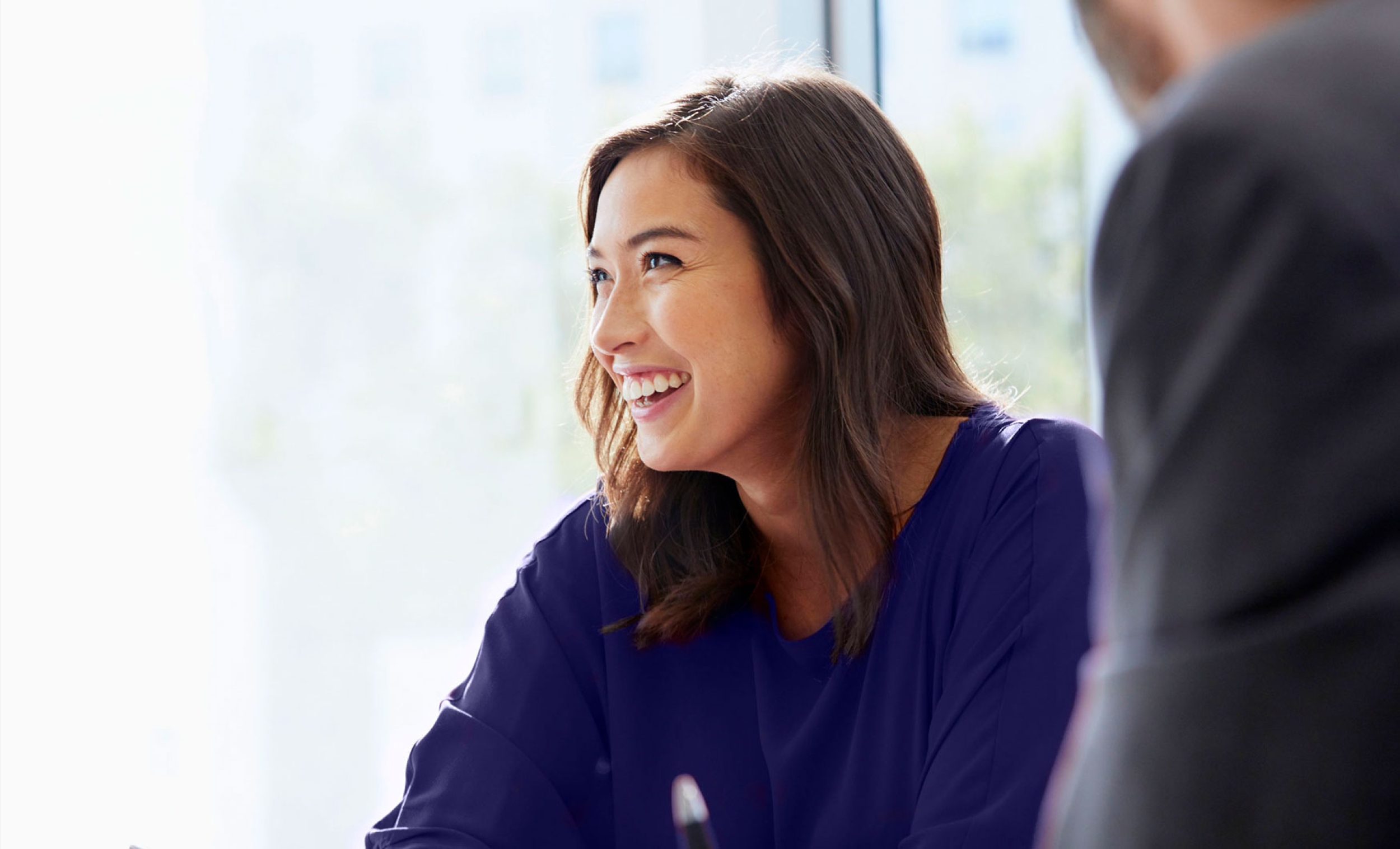 Business woman in a meeting with colleagues, smiling and wearing a purple top.