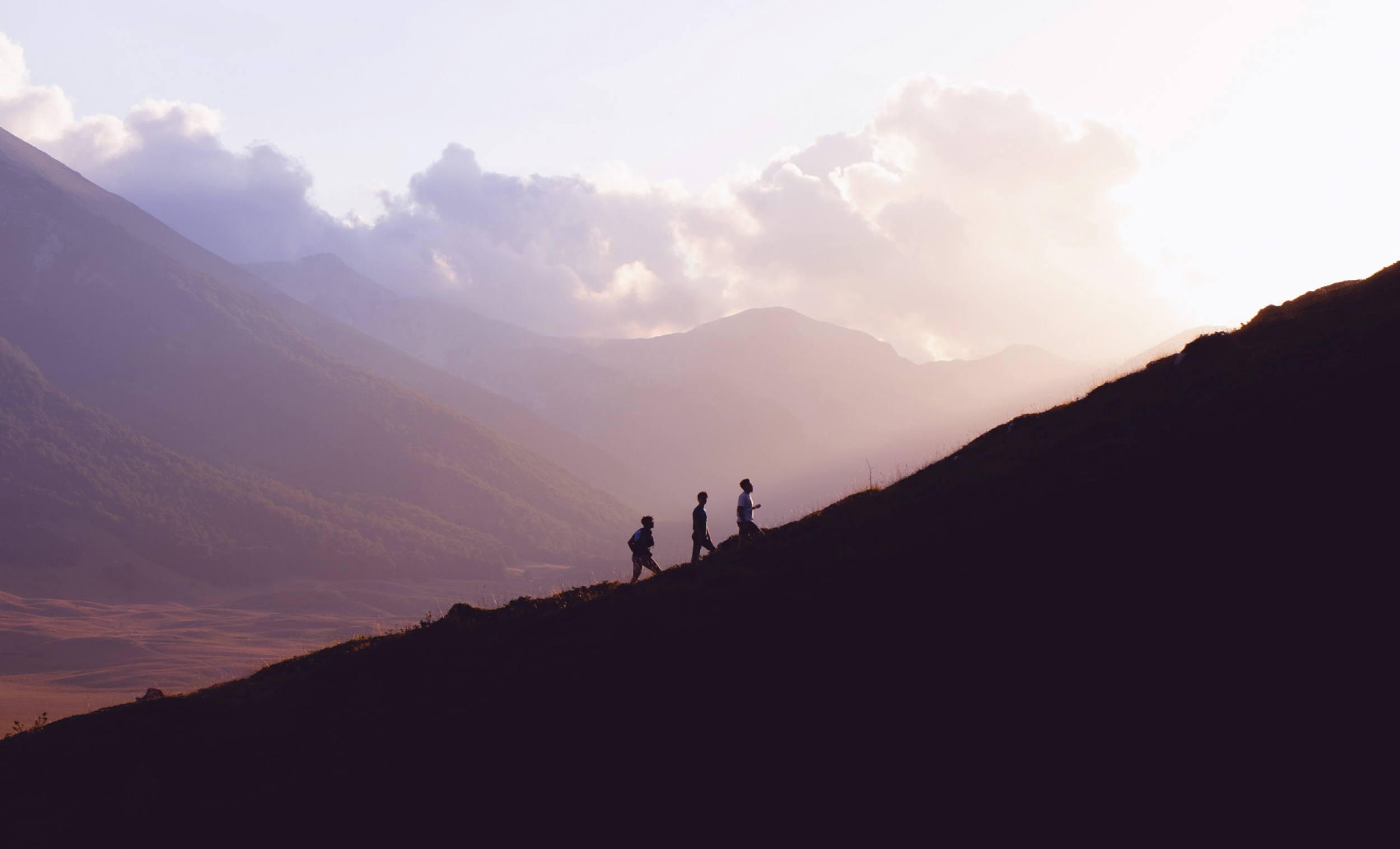 Three people seen from a distance while climbing a hill. A landscape of mountains and pink skies behind them.