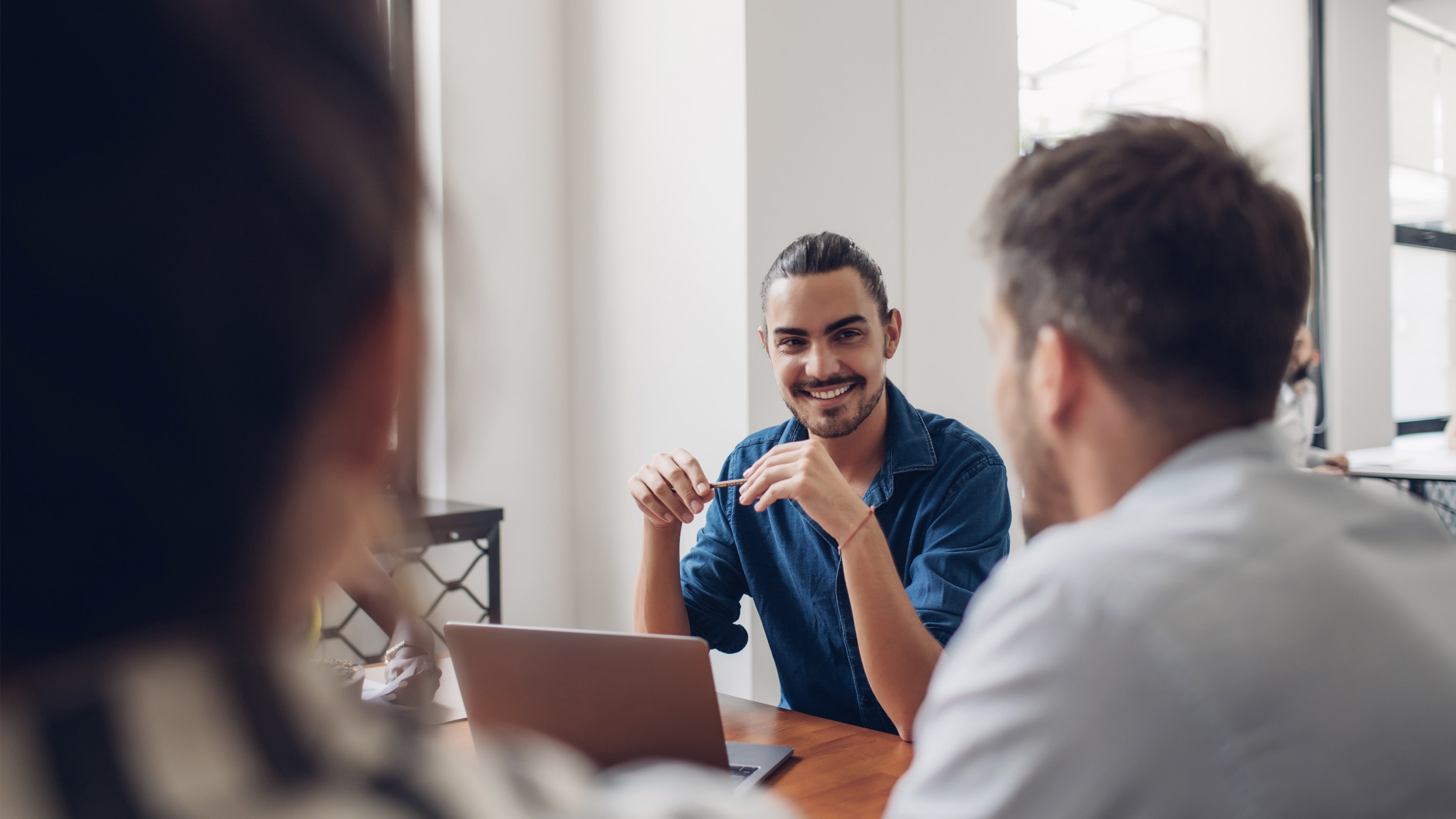 A meeting between a few office workers. One of them is in focus and is smiling at his coworker. There is a laptop in between them.