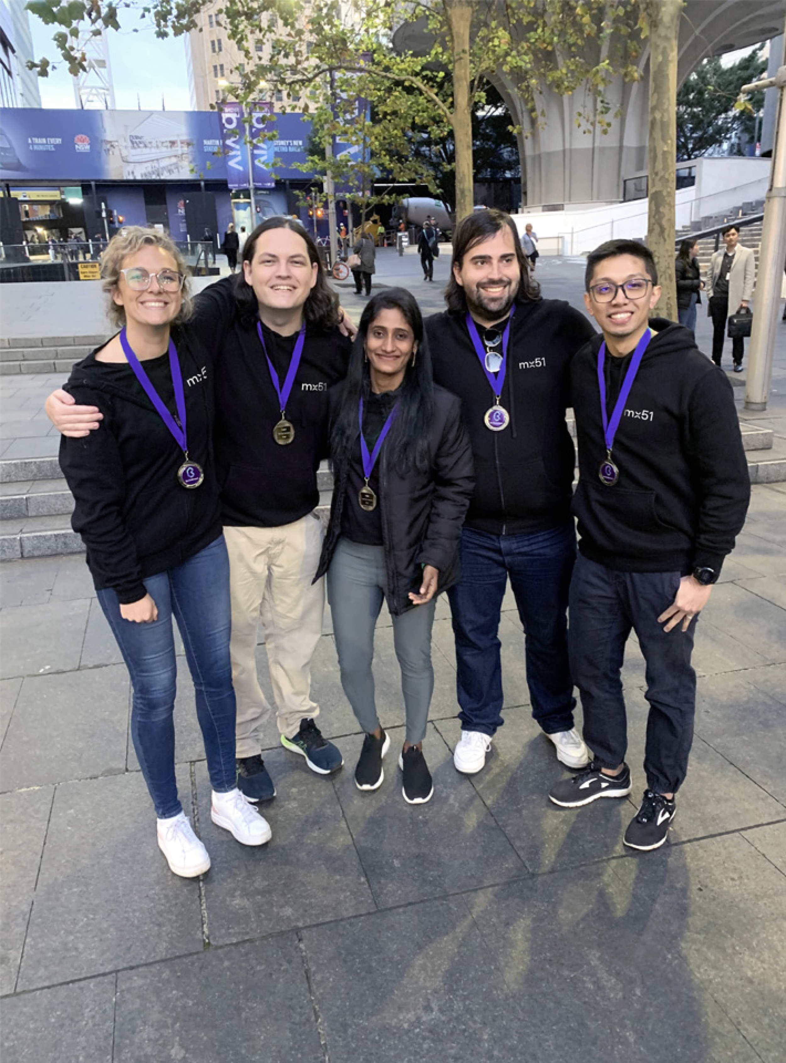 Snapshot of a small group of mx51 colleagues wearing mx51 jumpers and purple key-cords, embracing each other and smiling.