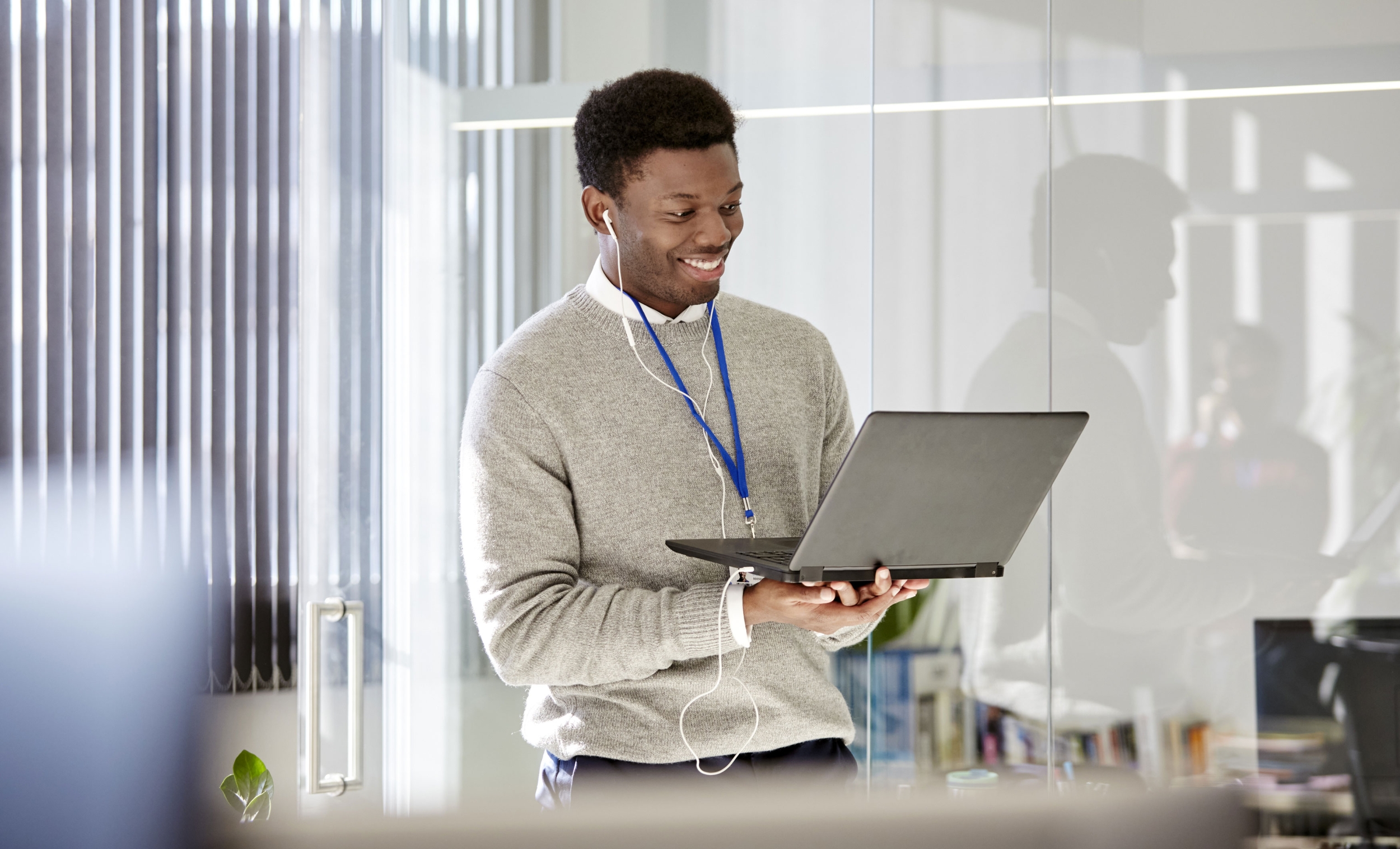 Young black business man wearing a grey jumper and a purple keycord in an office environment. He is holding up a laptop, wearing headphones and smiling at the laptop camera.