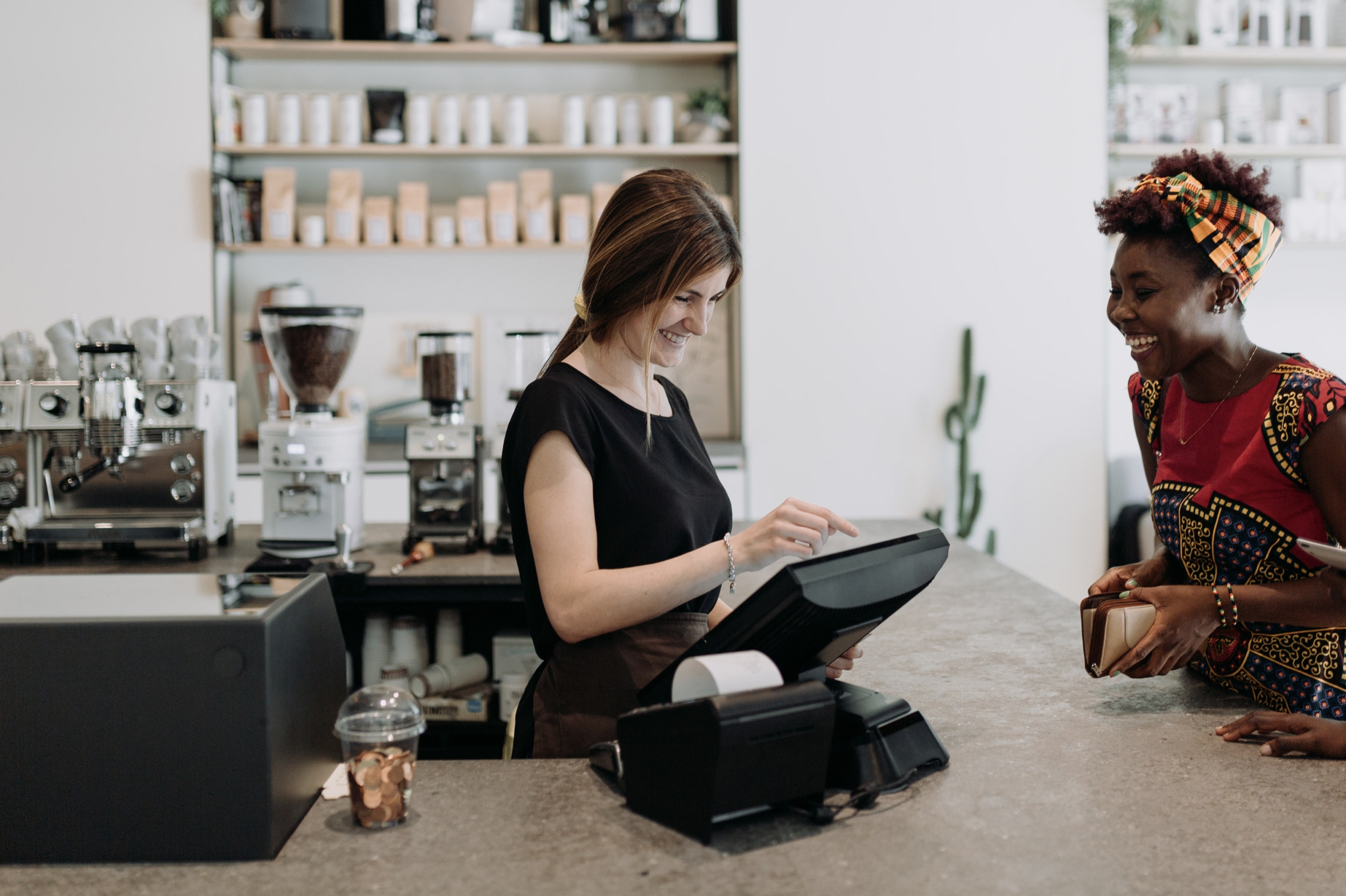 A smiling barista is using a computer with payments software to help a smiling customer pay.