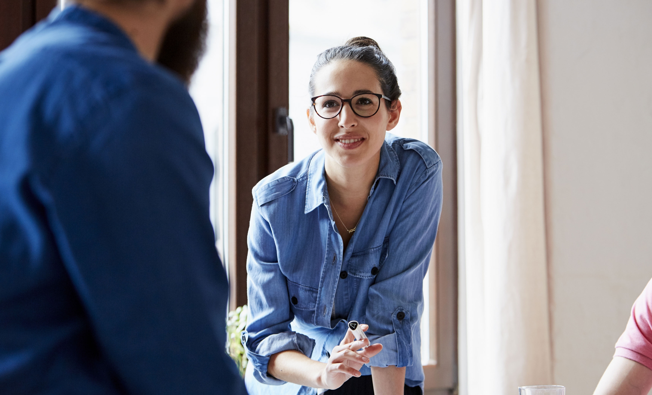 A group of colleagues are working together. Focus on a young female colleague wearing a denim shirt and glasses, holding a pen and smiling at her colleague.