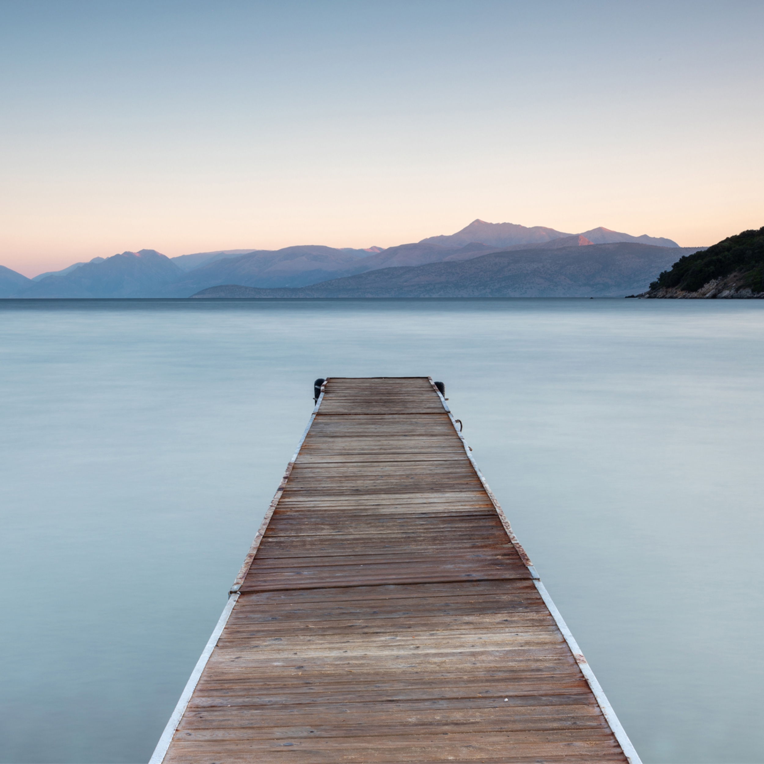 Photo of a tranquil environment of a lake and jetty with mountains in the background and a blue sky.