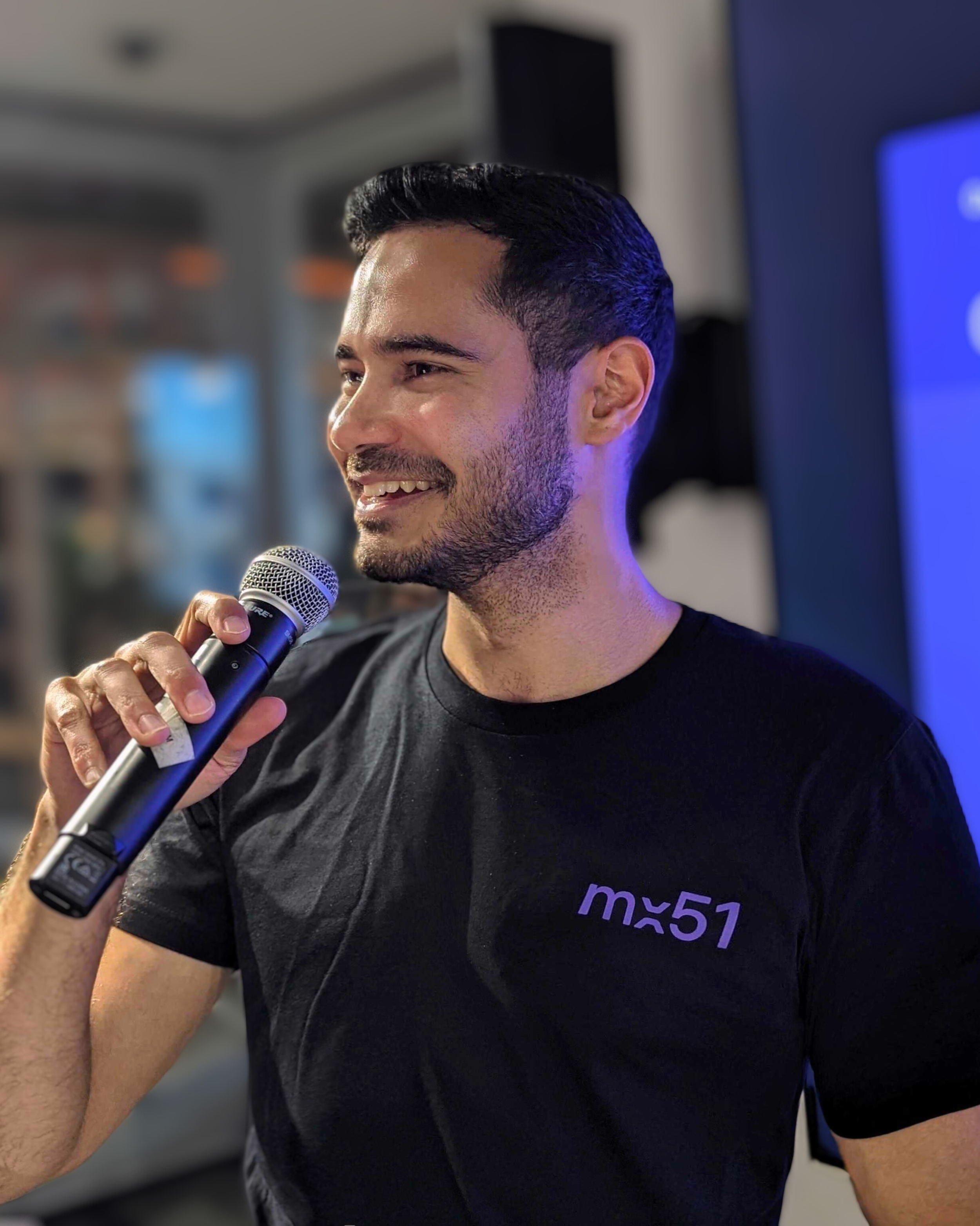 A dark-haired man in an mx51 t-shirt is smiling whilst holding a microphone in front of a presentation.