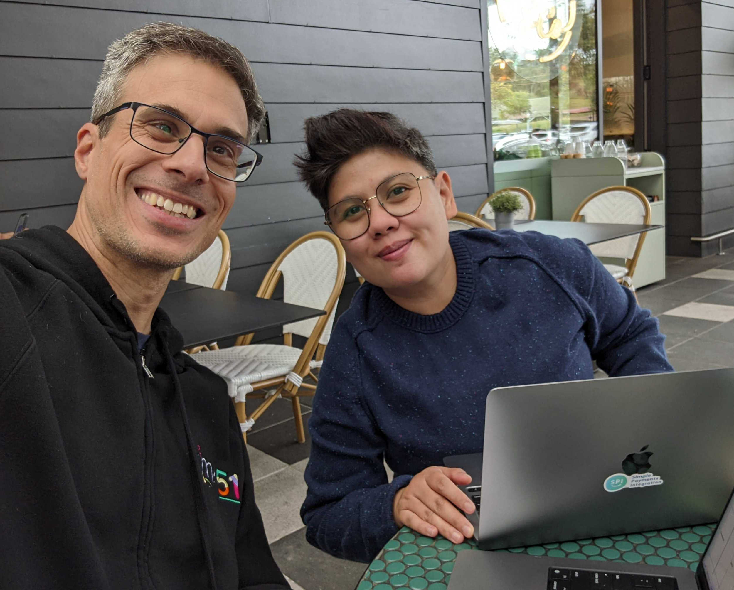 A selfie of two mx51 employees smiling for the camera, sitting at a cafe with their laptops.