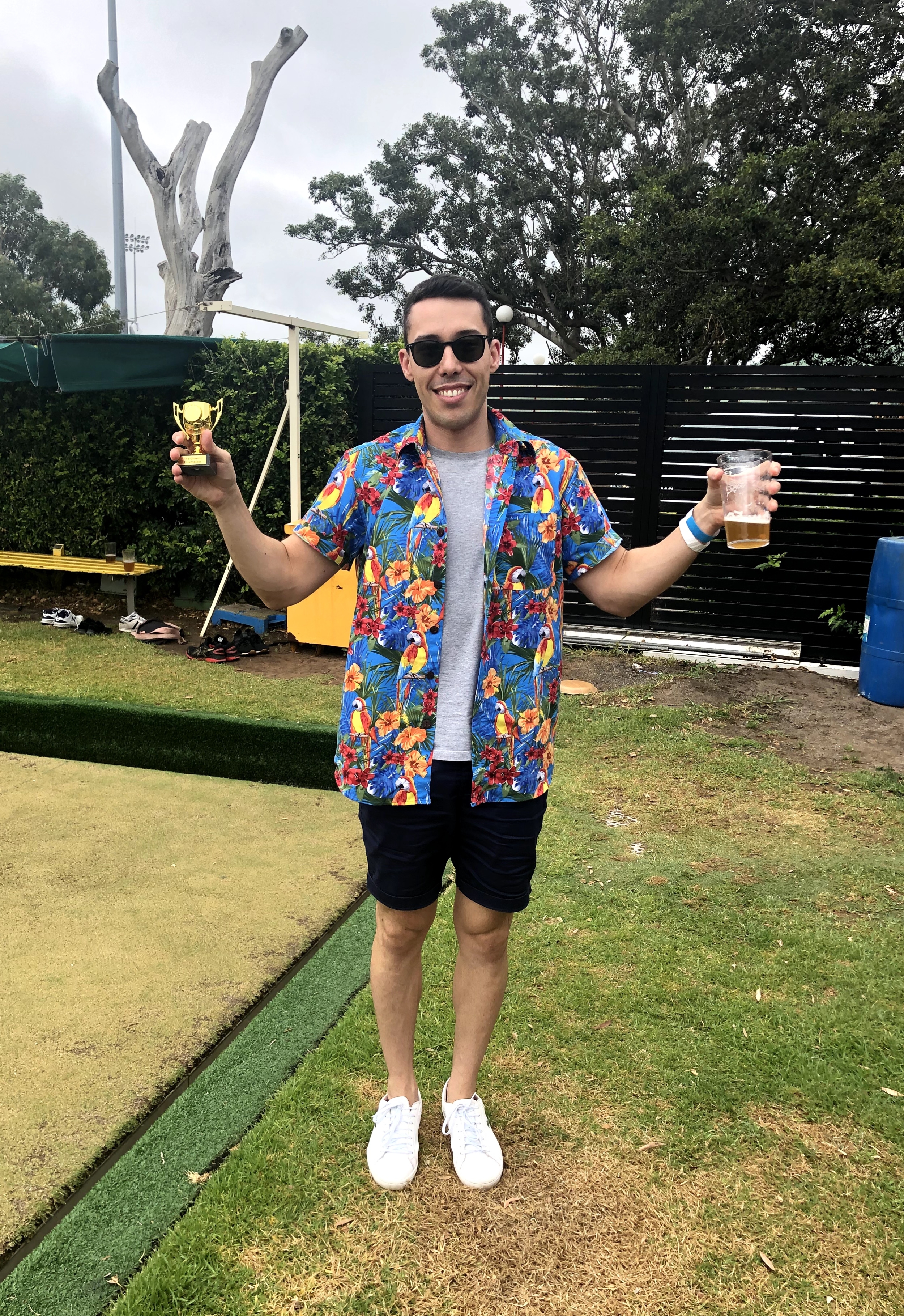 Snapshot of an mx51 employee holding a beer in one hand and a tiny trophy in the othe,r wearing sunglasses and a Hawaiian shirt, standing on the grass at a lawn bowls event.